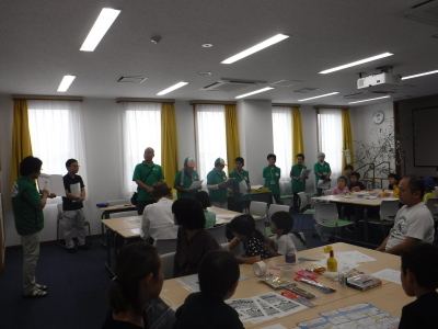 参加者達がグループ毎に分かれて席に着き、緑色のシャツを着た人の説明を聞いている写真