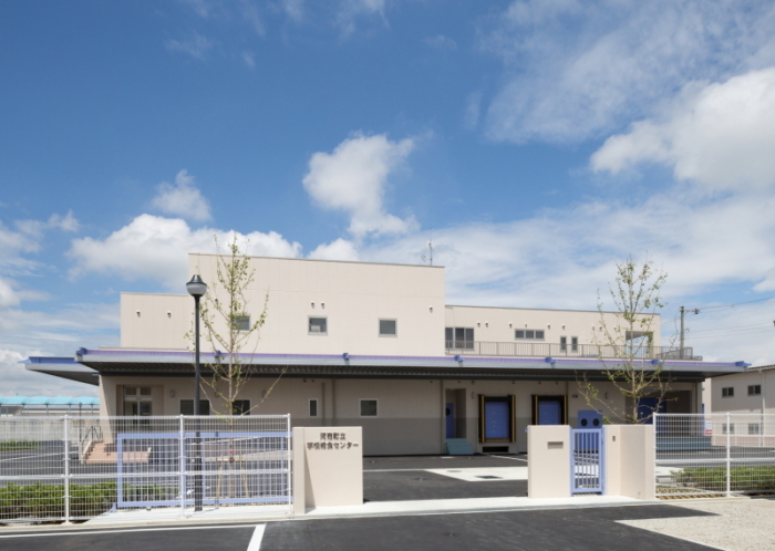 青空の下ベージュ色で2階建て、周囲をフェンスに囲まれ、入り口に門が設置してある給食センターの建物の外観写真