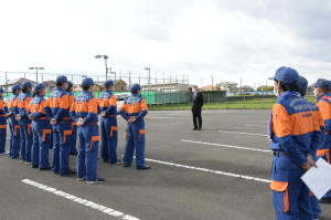広い駐車場にて、オレンジと青色の制服を着た消防団員たちが駐車場中央に立って町長の話を聞いている写真