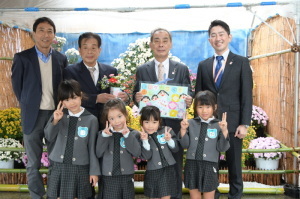 絵が描かれた紙を持っている森田町長、関係者の男性3名、ピースサインをしている園児4名の集合写真