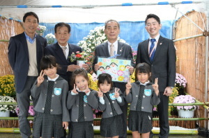 絵が描かれた紙を持っている森田町長、関係者の男性3名、ピースサインをしている園児4名の集合写真