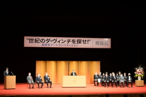 ステージ上に「世紀のダ・ヴィンチを探せ!」と書かれた釣り看板、中央に置かれた金屏風、ステージ両端にパイプ椅子に座っている関係者たち、中央の演題で話をしている男性の写真
