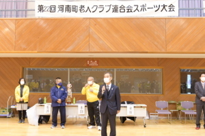 「第22回河南町老人クラブ連合会スポーツ大会」と書かれた横断幕が掲げられた会場でマイクを持ち話をしている町長の写真