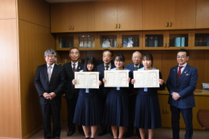 3名の女子中学生が賞状を持ち、スーツを着た関係者の方々5名と記念撮影をしている写真
