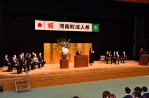 「祝 河南町成人祭」と書かれた吊り看板がステージ上に掲げられ、ステージ上には関係者たちが椅子に座っている成人祭の様子の写真