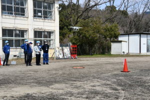 グラウンドに1台のドローンが飛んでおり、操縦技能資格講習会が行われている写真