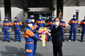 青とオレンジ色の制服を着て横一列に並んでいる消防団員たちの前で団員2名と森田町長が紅白のリボンがかけられた金色の大きなカギを一緒に持ち記念撮影をしている写真