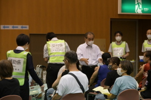 間隔をあけて椅子に座っているワクチン接種に来られた方々、黄色のゼッケンを付けた職員たちがいるワクチン接種会場の写真