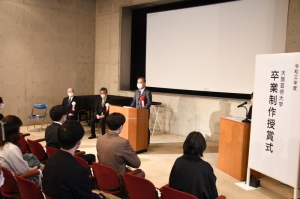 右側に「大阪芸術大学卒業制作授賞式」と書かれた看板があり、中央の演台で胸章を着けた町長が学生の皆さんへ向けて話をしている写真