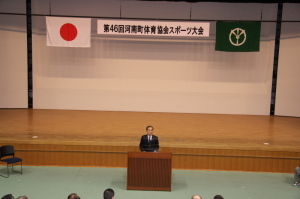 ステージ前に置かれた演題で話をしている男性を会場の高い位置から写したスポーツ大会開会式の写真