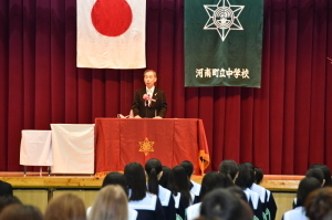 国旗と校章が掲げられたステージ上で町長が生徒たちに向かって話をしている様子の写真