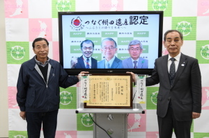 「つなぐ棚田遺産認定」と書かれ3名の男性の顔がモニターに映し出され、モニターの前に設置された台の上に置かれた賞状に手を添えて記念撮影をしている持尾棚田保存会の植田さんと森田町長の写真