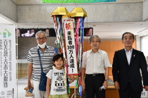 割れたくす玉から「祝 来館者300万人達成」と書かれた幕が出てきており、来館者の小西さん、高野さん、舘野館長、森田町長が記念撮影をしている写真