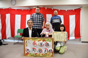 室内に張られた紅白幕の前で記念撮影をしている森田町長、丸茂さん、丸茂さんご家族の写真