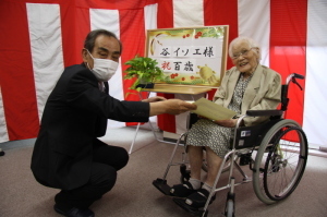 百歳長寿のお祝い訪問で車いすに座っている谷イソエさんに賞状を渡している町長の写真