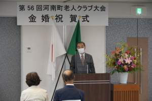 「第56回河南町老人クラブ大会 金婚祝賀式」と書かれた釣り看板が掲げられた会場で市長が演台に立ち話をしている様子の写真