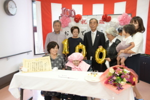 薄いピンク色をしたちゃんちゃんこと帽子を被った岩本さんと岩本さんご家族、森田町長が記念撮影をしている写真