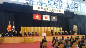広い会場のステージ上の両側に関係者たちが椅子に座り、中央でスーツを着た男性が立ち、演台の奥に立っている人のほうを見て話をしている大阪芸術大学入学式の写真