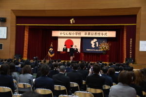 保護者や卒業生が椅子に座り、ステージ上で卒業証書授与式が行われている様子を体育館の後方から写した写真
