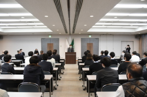 退任式に参加している職員の皆さんが長テーブルに2名ずつ座り、前方で男性が話をしている新田教育教育長退任式の様子を室内後方から写した写真
