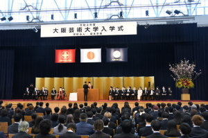 ステージ上に「大阪芸術大学入学式」と書かれた釣り看板が掲げられ、金屏風や大きな生け花がステージに設置され、多くの学生が入学式に参加している入学式会場全体を写した写真