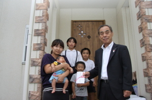 赤ちゃんを抱っこしているお母さんと、子供たち3人、新生児特別給付金を持っている町長が家の玄関前で記念撮影をしている写真