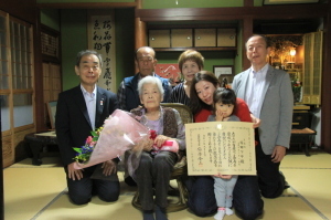 花束を持った高齢の女性と女性の家族、町長が畳の上に座って記念撮影をしている写真