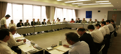 ロの字に設置された長机に参加者が座り、机上にある資料を見ながら会議をしている写真