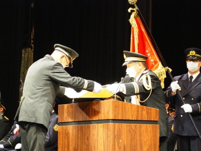 表彰旗を持った男性の前の壇上で、賞状の受け渡しをしている表彰式の写真