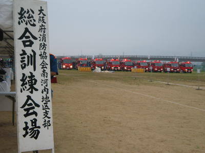大阪府消防協会南河内地区支部 総合訓練会場と書かれた案内板の奥に消防車両が並んでいる写真