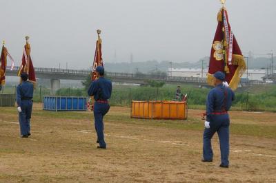 旗を持った消防団員3名が河原を歩いている後ろ姿を写した写真